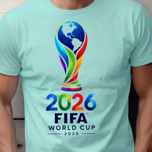 May include: A light mint green short-sleeve t-shirt featuring a colorful graphic of the FIFA World Cup 2026 trophy. The design includes the globe at the top, the year 2026 in large bold numbers, and the text FIFA WORLD CUP 2026 in black lettering.