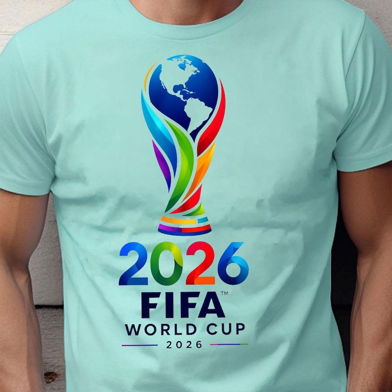 May include: A light mint green short-sleeve t-shirt featuring a colorful graphic of the FIFA World Cup 2026 trophy. The design includes the globe at the top, the year 2026 in large bold numbers, and the text FIFA WORLD CUP 2026 in black lettering.