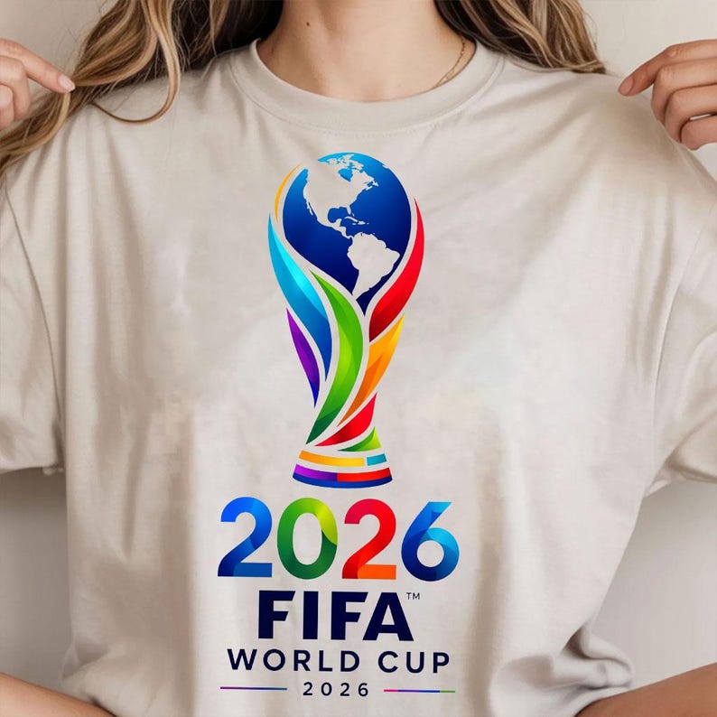 May include: A beige t-shirt featuring a colorful graphic of the FIFA World Cup 2026 trophy, which incorporates a globe design. Below the trophy, the text reads 2026 FIFA WORLD CUP 2026 in bold blue and black lettering with a small horizontal line.