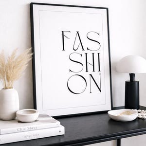 Könnte beinhalten: Schwarz gerahmter Druck mit dem Wort "FASHION" in minimalistischer Schrift. Auf einem schwarzen Tisch stehen eine weiße Vase mit Trockenblumen, eine kleine Schale, Bücher und eine moderne Lampe.