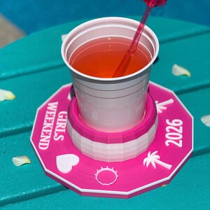 Könnte beinhalten: Ein weißer Plastikbecher gefüllt mit einem orangenen Getränk, auf einem pinkfarbenen Untersetzer. Der Untersetzer trägt die Aufschrift "GIRLS WEEKEND 2026" und hat dekorative Elemente. Ein pinkfarbener Strohhalm mit blütenförmigem Aufsatz steckt im Getränk.