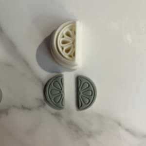 May include: A white and gray citrus-shaped cookie cutter and two gray citrus-shaped cutouts. The cookie cutter is a half-circle with a detailed interior design. The cutouts are also half-circles with a similar design.