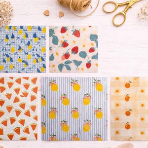 May include: A collection of six patterned beeswax food wraps. Designs include lemons, strawberries, watermelon slices, and oranges. The wraps are arranged on a white wooden surface with floral accents and gold scissors.