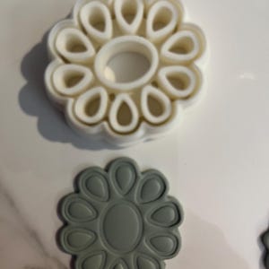 May include: Two flower-shaped cookie cutters. One is white, with a central ring and petal-shaped cutouts. The other is gray, with a similar design. These tools are ideal for baking and crafting.