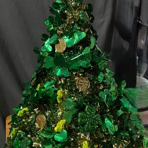 Può includere: Un festoso albero di San Patrizio, adornato con trifogli verdi, monete d'oro e accenti glitterati. L'albero è ricoperto di orpelli verdi e ha un gruppo di trifogli verdi scintillanti in cima.