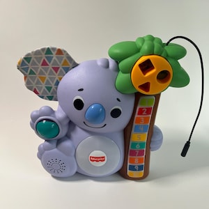 Pode incluir: Um brinquedo interativo Fisher-Price de coala em tons de roxo, verde e castanho. O coala tem um nariz azul, uma orelha estampada e uma palmeira com números. O brinquedo tem um altifalante e um botão com o logótipo Fisher-Price.