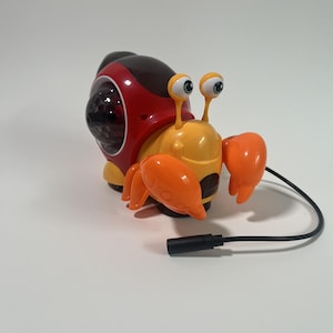 May include: A yellow and orange toy crab with a red and black shell. The crab has large, googly eyes and orange claws. A black cord extends from the crab's body. The toy is designed to look like a crab and a snail.