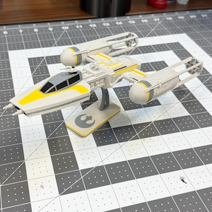 May include: A white and yellow Y-wing starfighter model with a black cockpit canopy, mounted on a gray display stand featuring a Rebel Alliance symbol. The spacecraft has two long engine nacelles and a central fuselage with yellow accent stripes.