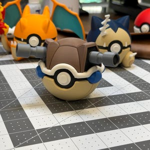 May include: A collection of colorful, 3D-printed Pokémon figures. The foreground features a brown and tan figure with two gray tubes extending from its sides. Other figures include orange, blue, and red characters.