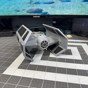 May include: A gray and black model of a TIE fighter spacecraft, a fictional starship from the Star Wars franchise. The model has detailed wings and a central cockpit section. The spacecraft is set against a background of a computer monitor.