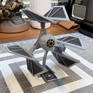 May include: A gray TIE defender model kit featuring a central spherical cockpit with a circular viewport, three distinct wing panels with black grid detailing, and a display stand with an Imperial logo emblem on the base.