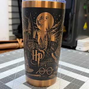 May include: A black stainless steel tumbler with a copper-colored rim and base. The exterior features a detailed laser-etched design in copper, depicting a castle, a full moon, the HP logo, round glasses, and a lightning bolt symbol.