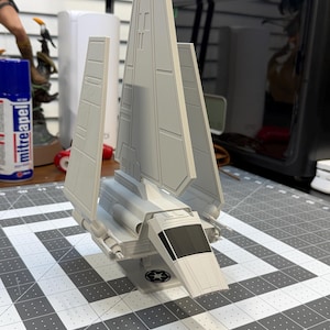 May include: A light gray model of a spacecraft with angled wings and a cockpit. The model is on a gray and white grid surface. The spacecraft has a detailed design and is likely a collectible item.