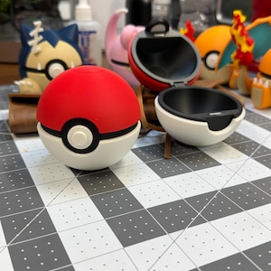 May include: A red and white Poké Ball, with a black center circle, sits open next to a closed one. Other Pokémon figures are in the background. The open ball reveals a black interior. The balls are on a gray and white grid.