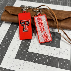 May include: Two red rectangular keychains. One features a black and white "T" logo, the other has the words "WRECK 'EM" in red over iridescent glitter. Both have silver key rings and are displayed on a brown leather wallet.