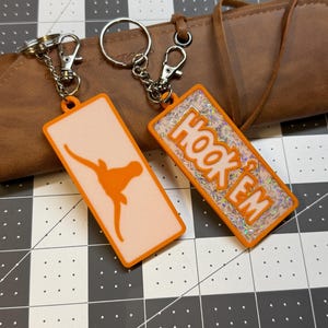 May include: Two rectangular keychains with orange borders and silver hardware. One keychain features an orange Longhorn silhouette on a peach background. The other has the words "HOOK 'EM" in orange with a glittery background. Both have a clasp and chain.