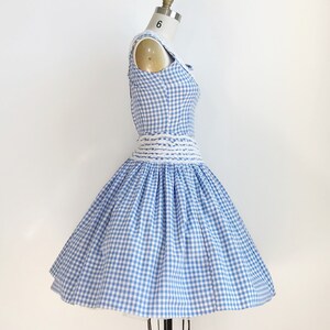 May include: A vintage-style dress in a blue and white gingham pattern. The sleeveless dress features a fitted bodice, a cinched waist with a white woven belt, and a flared skirt. The dress is displayed on a dress form.
