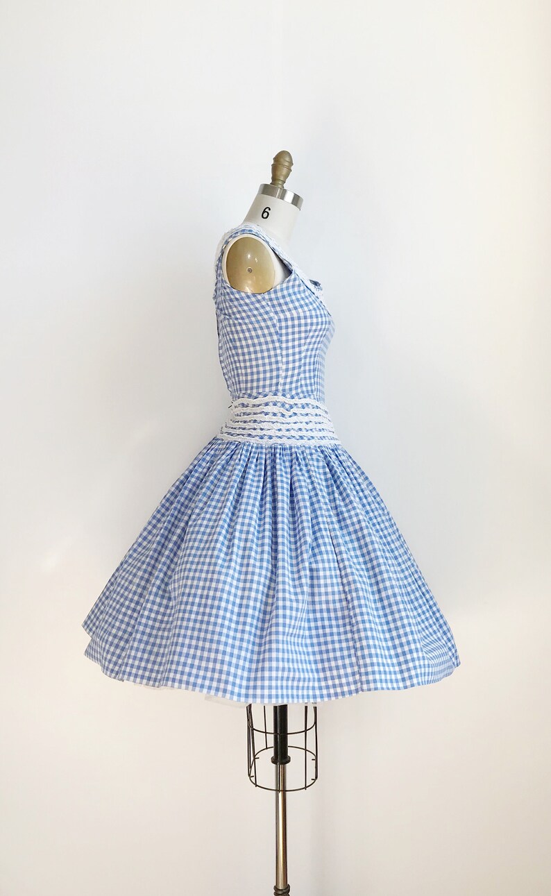 May include: A vintage-style dress in a blue and white gingham pattern. The sleeveless dress features a fitted bodice, a cinched waist with a white woven belt, and a flared skirt. The dress is displayed on a dress form.