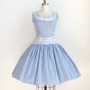 May include: A sleeveless blue and white gingham dress with a full skirt, displayed on a mannequin. The dress features a white lace collar and a white band at the waist. The skirt is gathered and flares out, creating a classic vintage silhouette.