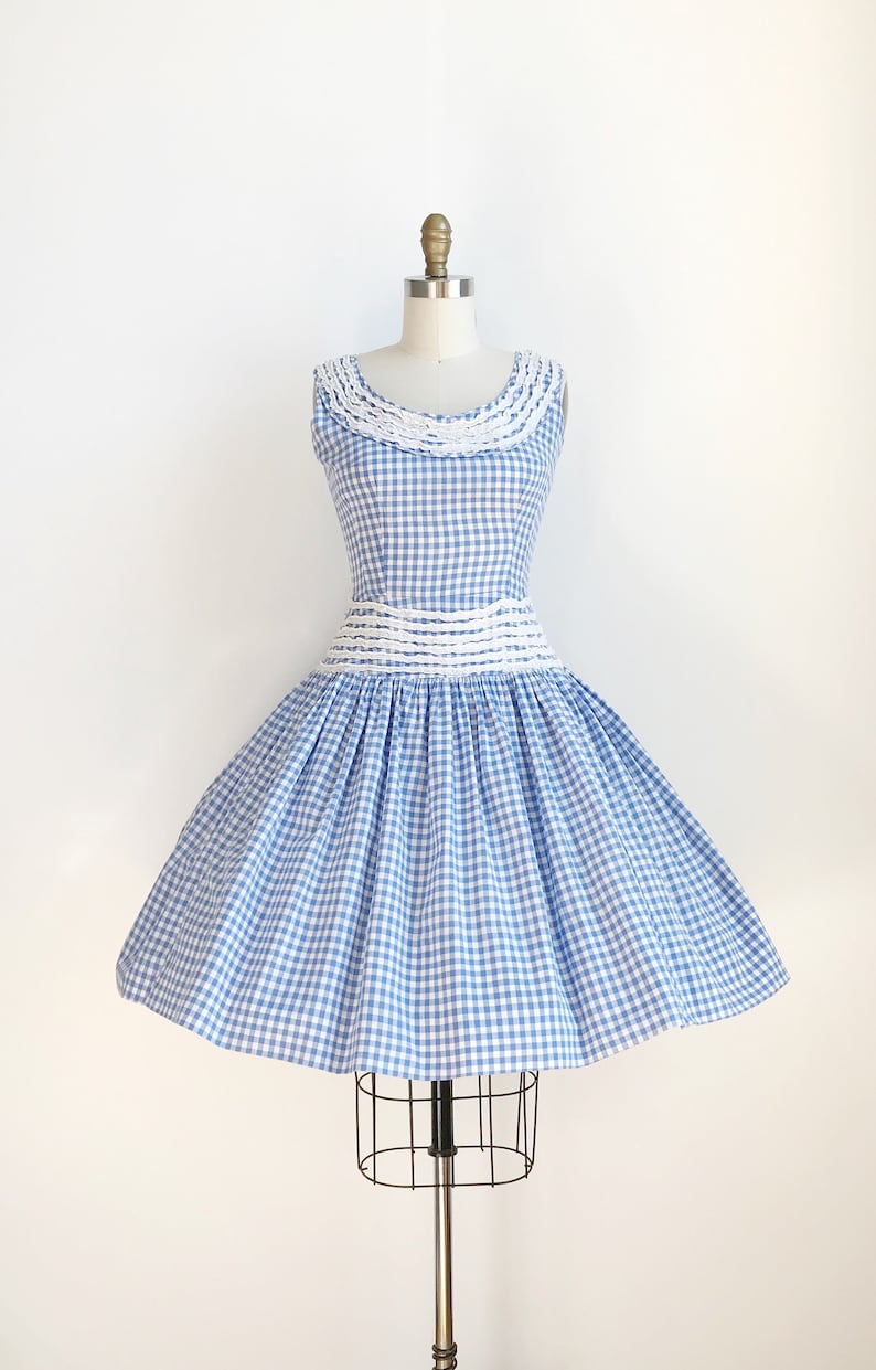 May include: A sleeveless blue and white gingham dress with a full skirt, displayed on a mannequin. The dress features a white lace collar and a white band at the waist. The skirt is gathered and flares out, creating a classic vintage silhouette.