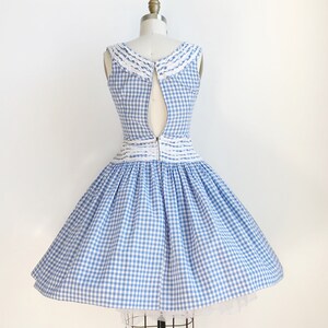May include: A vintage-style blue and white gingham dress on a mannequin. The sleeveless dress features a white lace-trimmed collar and waist detail. The skirt is full and flared, and the dress is zipped up the back.