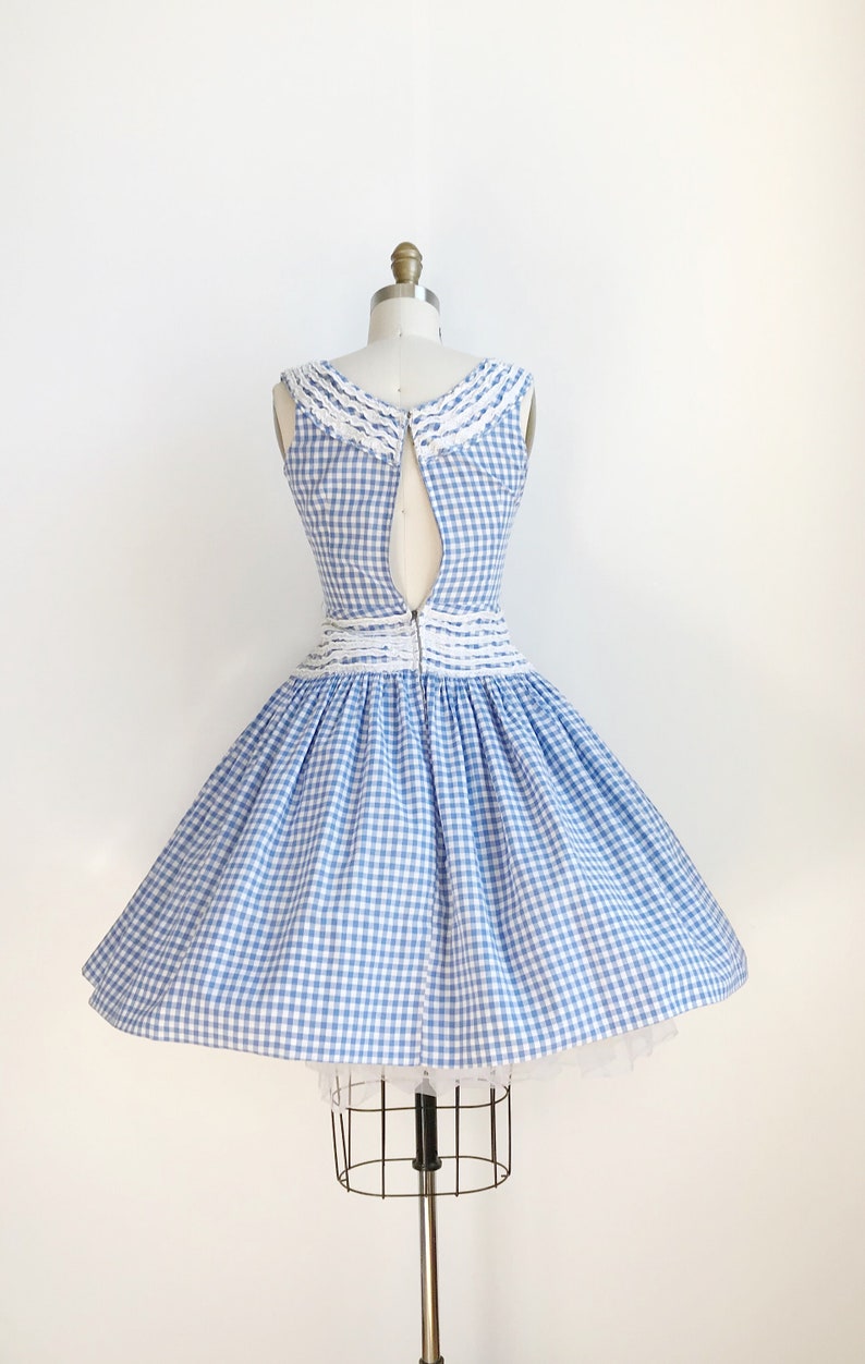 May include: A vintage-style blue and white gingham dress on a mannequin. The sleeveless dress features a white lace-trimmed collar and waist detail. The skirt is full and flared, and the dress is zipped up the back.