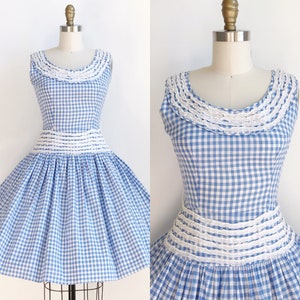 May include: A sleeveless blue and white gingham dress with a white lace trim around the neckline and waist. The dress has a full skirt and is displayed on a mannequin.