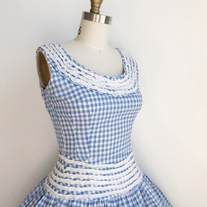 May include: A vintage blue and white gingham dress with a white lace trim around the neckline and waist. The dress is sleeveless and has a full skirt, displayed on a mannequin.