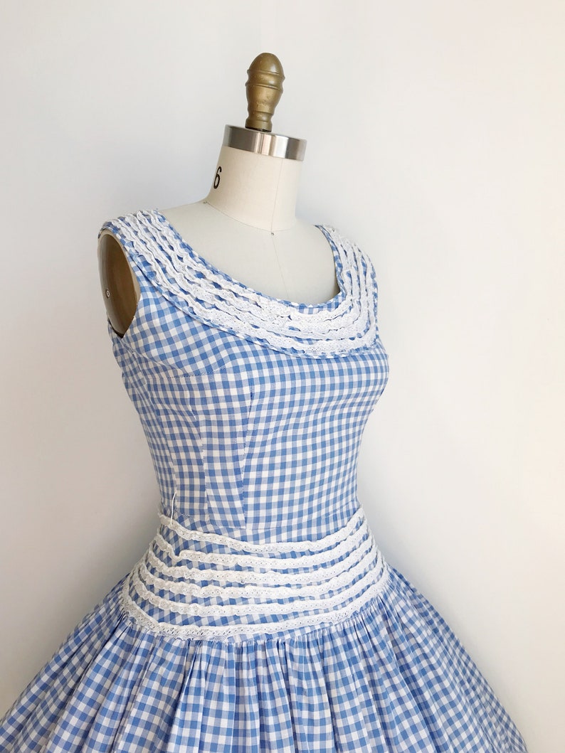May include: A vintage blue and white gingham dress with a white lace trim around the neckline and waist. The dress is sleeveless and has a full skirt, displayed on a mannequin.
