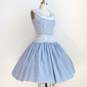 May include: A vintage blue and white gingham dress on a mannequin. The sleeveless dress features a fitted bodice with a white crocheted neckline and waist detail. The skirt is full and flared, creating a classic silhouette. The dress is a light blue and white check pattern.