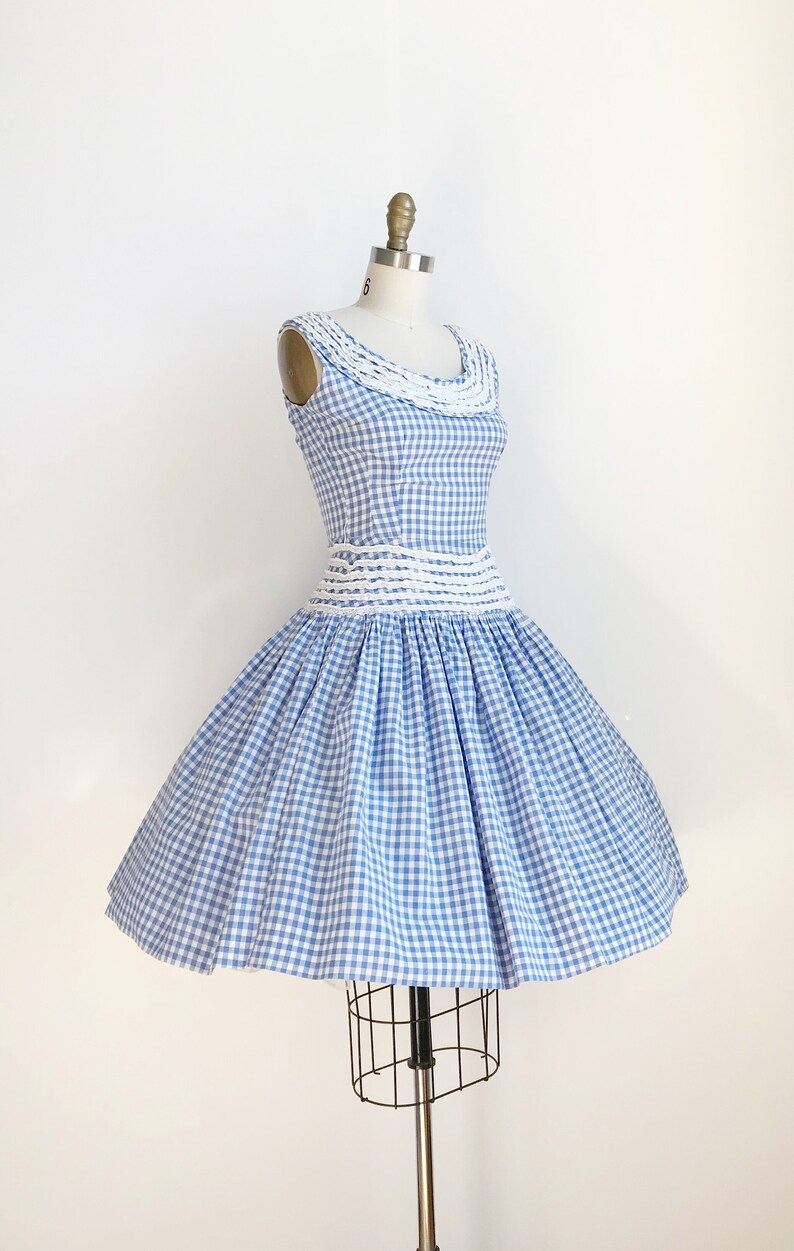 May include: A vintage blue and white gingham dress on a mannequin. The sleeveless dress features a fitted bodice with a white crocheted neckline and waist detail. The skirt is full and flared, creating a classic silhouette. The dress is a light blue and white check pattern.