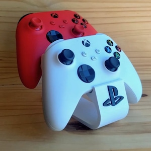 May include: A red Xbox controller is stacked on top of a white PlayStation controller, which is resting on a white stand with a black PlayStation logo. The controllers have black buttons and joysticks.
