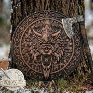 Op de afbeelding: Een rond houten schild met een centraal reliëf van een grommende wolvenkop, omringd door ingewikkelde Keltische knoopmotieven en de tekst FENRIR, vergezeld van een bijpassende strijdbijl met een gedetailleerd metalen blad en handvat.