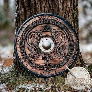 May include: A circular wooden shield featuring a central metal boss, intricate carvings of two dragons, a triquetra knot, and a hammer symbol. The outer rim displays the text WELCOME and WALHALLA with runic-style lettering and metal rivets.