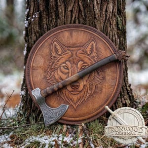 Op de afbeelding: Een rond bruin lederen schild met een reliëf van een wolvenkop, gecombineerd met een metalen bijl met een ingewikkeld gegraveerd patroon op het blad en een houten handvat. Een klein rond embleem met de tekst Ironwood Arsenal is inbegrepen.