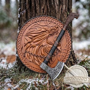 May include: A circular wooden shield featuring a carved wolf head design and a metal chain detail, paired with a battle axe that has a wolf-shaped pommel, a leather-wrapped handle, and an engraved blade. A small circular logo reads IRONWOOD ARSENAL.
