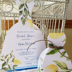 Puede incluir: Invitaci&oacute;n y caja de regalo para despedida de soltera blancas, ambas con forma de vestido, adornadas con limones y hojas verdes en acuarela. La invitaci&oacute;n incluye los detalles del evento. La caja de regalo est&aacute; atada con hilo. Las palabras "Beautifully Inviting By Beata" est&aacute;n escritas en la parte inferior.