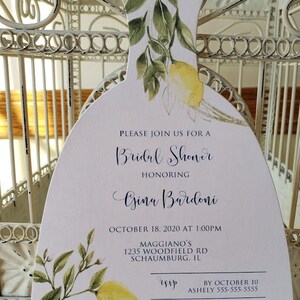 Puede incluir: Una invitaci&oacute;n blanca en forma de vestido para una despedida de soltera, adornada con limones y vegetaci&oacute;n en acuarela. La invitaci&oacute;n incluye el texto "Bridal Shower" y los detalles del evento, incluyendo la fecha, hora y lugar.