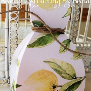 Puede incluir: Una caja de regalo blanca con un dise&ntilde;o de lim&oacute;n y hojas, atada con hilo. La caja presenta limones amarillos y hojas verdes sobre un fondo rosa claro. El texto en la caja dice "Beautifully Inviting By Beata".