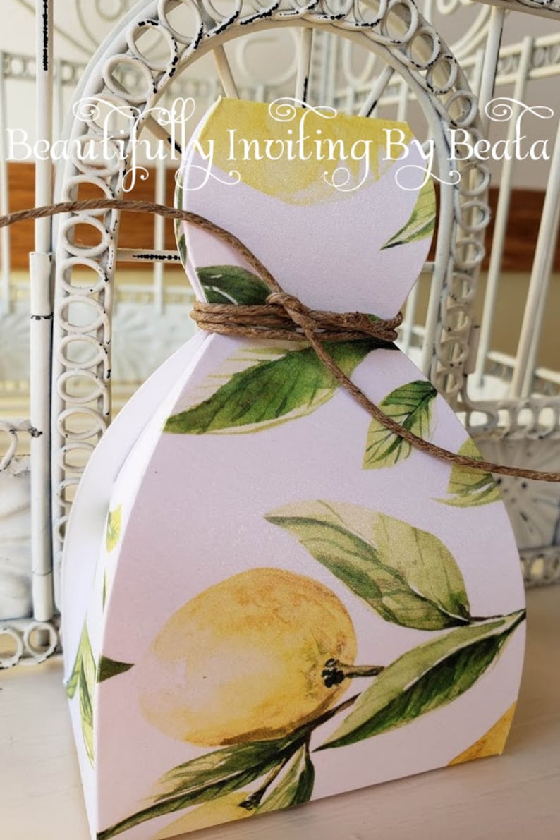 Puede incluir: Una caja de regalo blanca con un dise&ntilde;o de lim&oacute;n y hojas, atada con hilo. La caja presenta limones amarillos y hojas verdes sobre un fondo rosa claro. El texto en la caja dice "Beautifully Inviting By Beata".