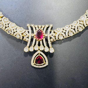 May include: A gold-toned necklace and matching earrings set. The necklace features a central pendant with a large, red gemstone and smaller white stones. The earrings mirror the pendant design, with red and white stones. The jewelry is displayed on a black mannequin.