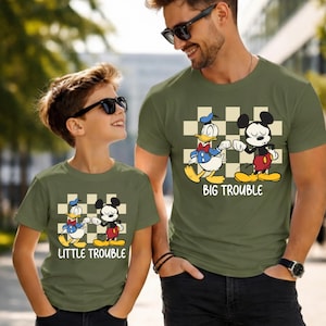 May include: Olive green t-shirts with a cartoon graphic of Donald Duck and Mickey Mouse. The shirts have the text "BIG TROUBLE" and "LITTLE TROUBLE" in white. The shirts are worn by a father and child.