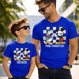 May include: Two matching blue t-shirts featuring Donald Duck and Mickey Mouse characters. One shirt says "Main Character" and the other says "Sidekick". The shirts have a checkered background and are worn by a father and son.