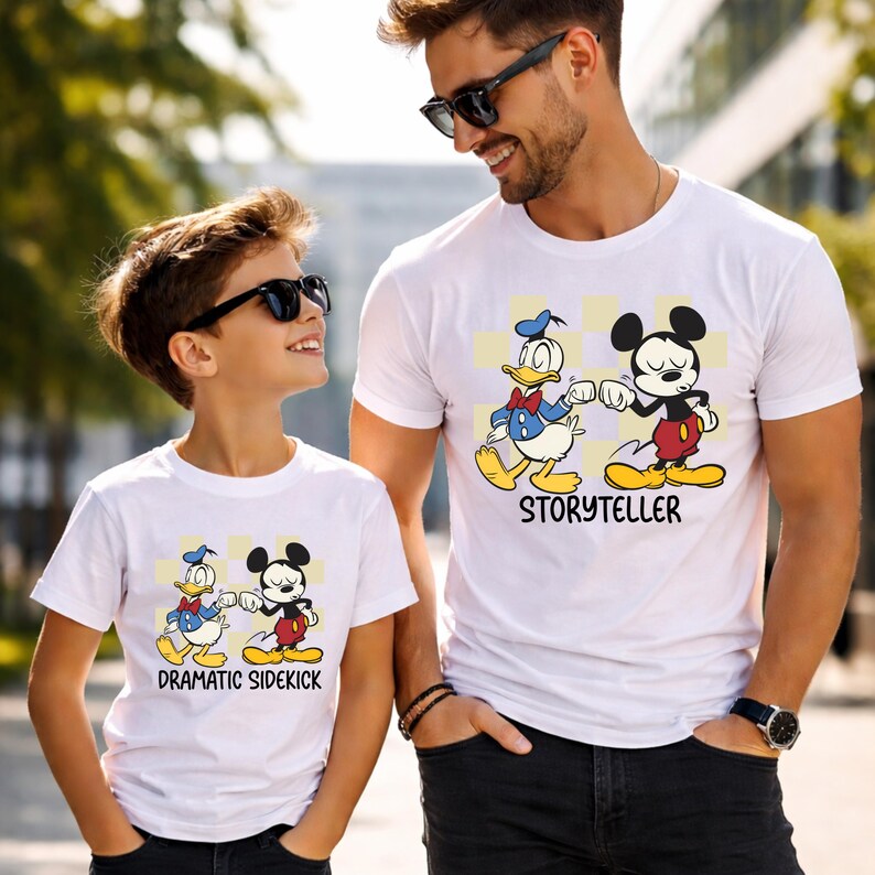 May include: White t-shirts featuring Donald Duck and Mickey Mouse fist-bumping. The shirt on the right says "Storyteller" and the shirt on the left says "Dramatic Sidekick". The shirts are worn by a man and a child.