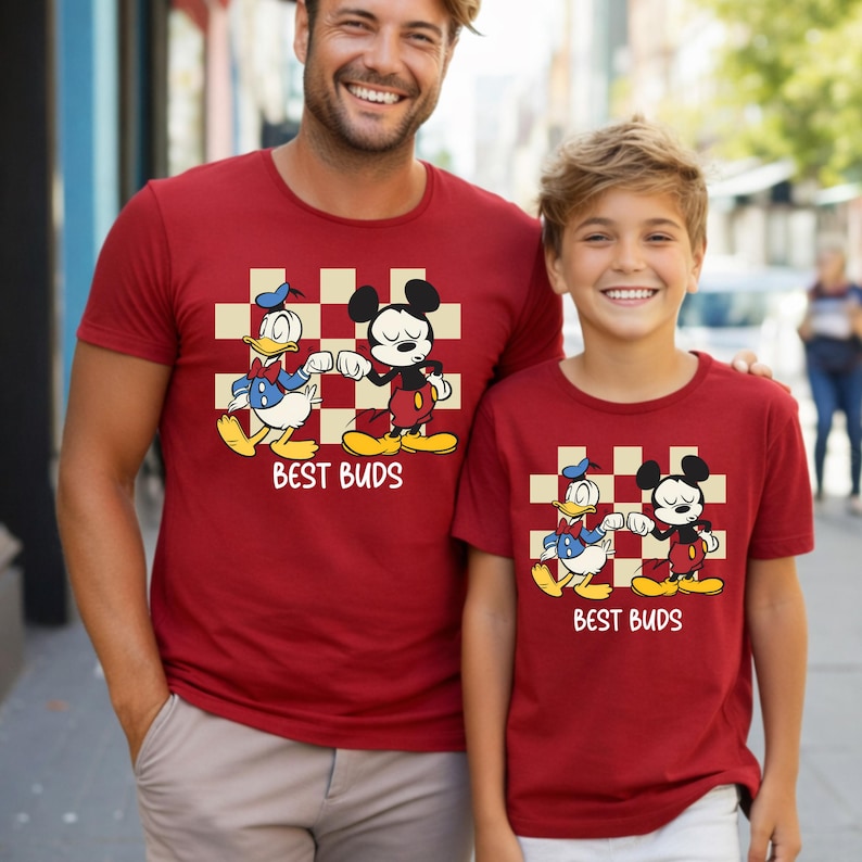May include: Two matching red t-shirts with a cartoon graphic of Mickey Mouse and Donald Duck bumping fists, set against a checkered background. The text "BEST BUDS" is below the graphic. The shirts are worn by a man and a child.