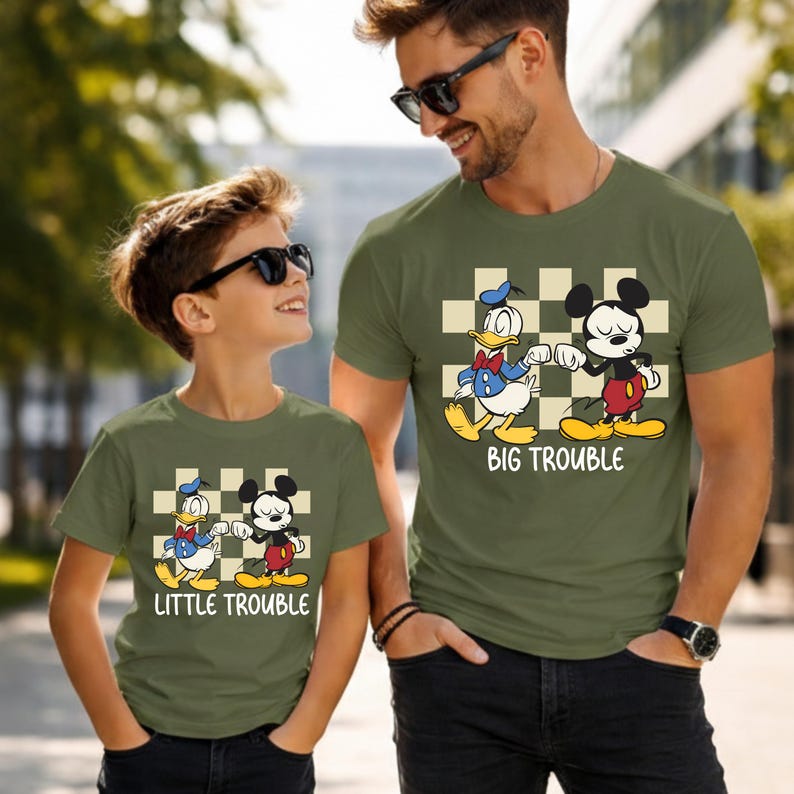 May include: Olive green t-shirts with a cartoon graphic of Donald Duck and Mickey Mouse. The shirts have the text "BIG TROUBLE" and "LITTLE TROUBLE" in white. The shirts are worn by a father and child.