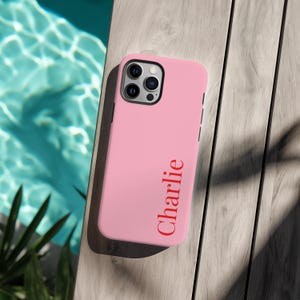 Könnte beinhalten: Eine rosa Handyhülle mit dem Namen "Charlie" in Rot aufgedruckt. Die Hülle wird auf einer Holzoberfläche neben einem Pool gezeigt. Die Handyhülle hat eine Kamera und Lautstärketasten.
