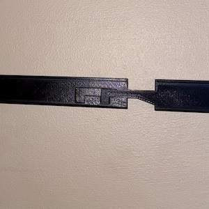 May include: A black, rectangular, 3D-printed object with a textured surface. It has a unique interlocking design with a central section and two extending arms. The object has pre-drilled holes for mounting hardware.