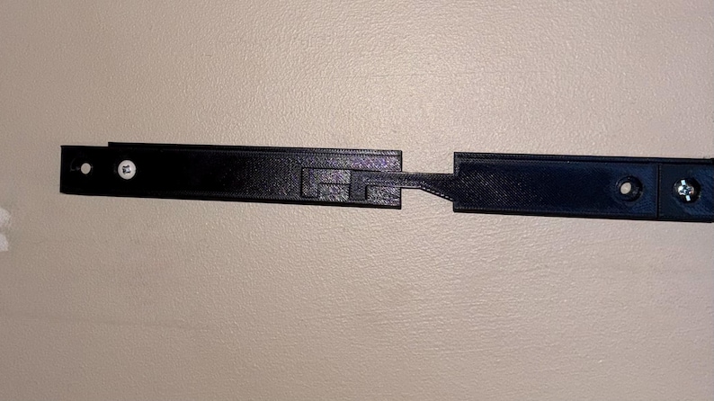 May include: A black, rectangular, 3D-printed object with a textured surface. It has a unique interlocking design with a central section and two extending arms. The object has pre-drilled holes for mounting hardware.
