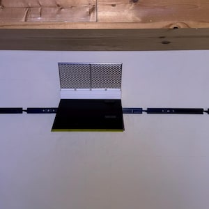 May include: A black and silver range hood with a rectangular design. The hood has a mesh screen and a black panel. It is mounted on a white wall with black support bars extending to the sides.